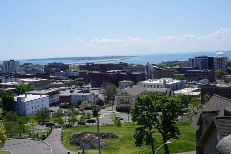 A cityscape view of Portland, Maine, showing buildings, green trees, and parks in the foreground, with the coastline and ocean visible in the distance under a clear blue sky.