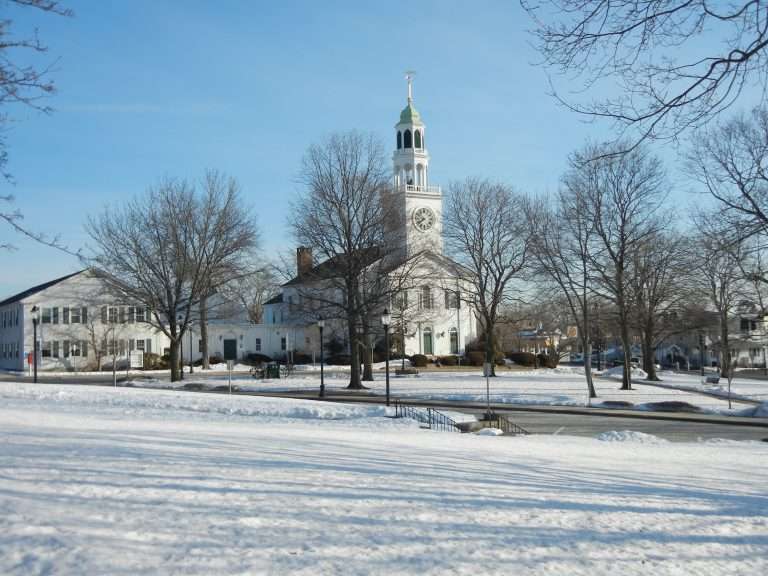 A white church with a tall clock tower stands behind leafless trees in a snowy Reading, MA park under a clear blue sky. Shadows from the trees stretch across the snow-covered ground.