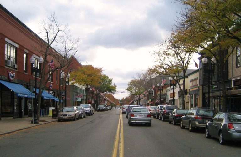 A two-lane street in the small town of Melrose, MA is lined with parked cars, storefronts, and trees with autumn leaves under a cloudy sky. Buildings display American flags, and the street appears calm and quiet.