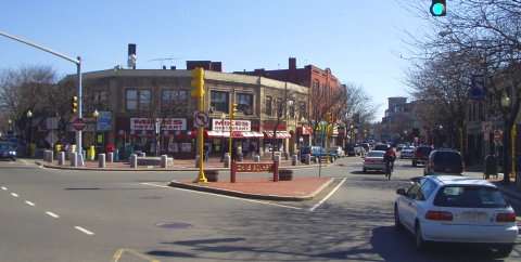 A city street scene in Somerville, MA, with cars driving and parked, storefronts with red awnings, and pedestrians on sidewalks. The sky is clear, a traffic light is green, trees line the street, and a yellow bench sits in a small plaza.