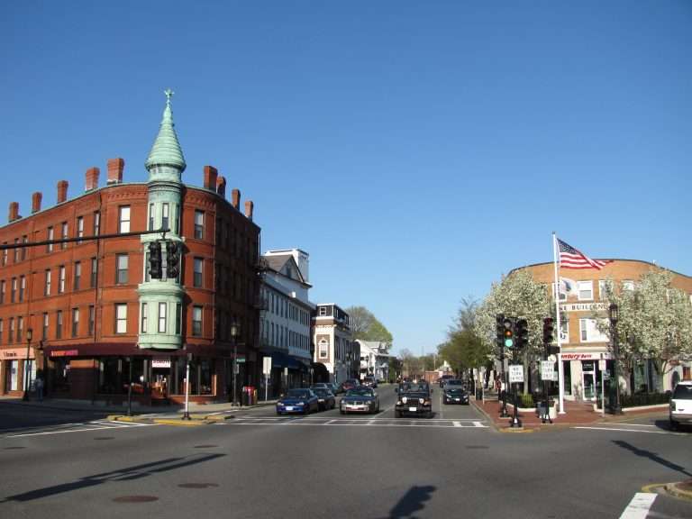 A small town intersection in Medford, MA with brick buildings, a green turret, shops, cars at a stoplight, and a clear blue sky. An American flag waves atop a building on the right, and trees with white blossoms line the street.