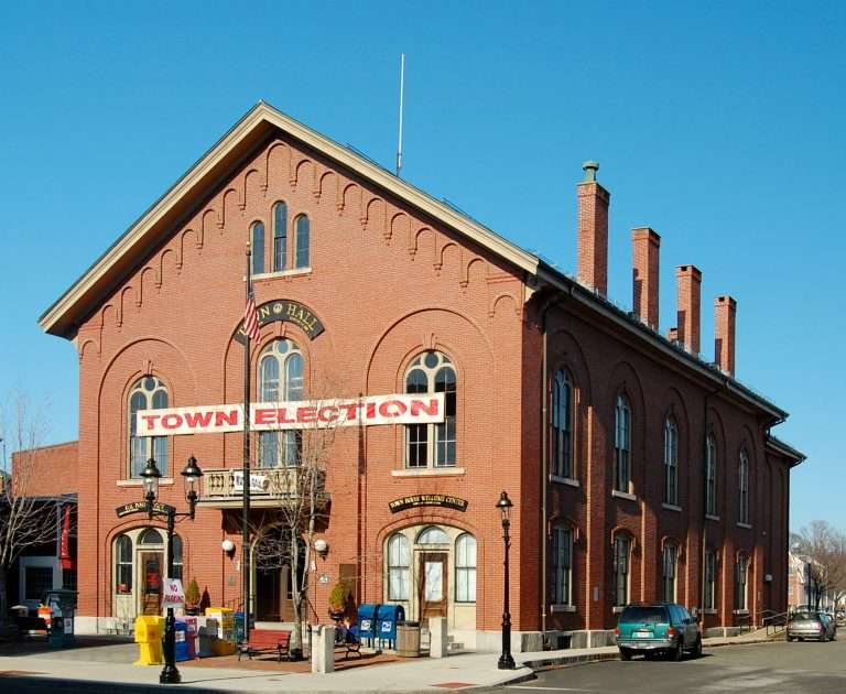 A historic red-brick town hall with arched windows displays a large Town Election banner above the entrance; American flag and lampposts are in front, and cars are parked along the street under a clear blue sky.
