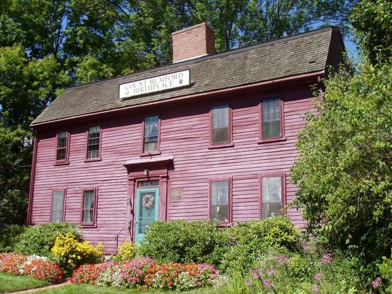 A two-story, red wooden colonial house with a sign reading Count Rumford Birthplace above the door, surrounded by green plants and colorful flowers, with trees in the background.
