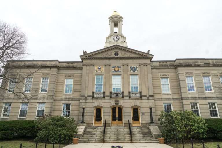 A historic beige stone building with a central clock tower, large windows, and decorative crests above two main wooden doors. Steps and bushes lead up to the entrance. The sky is overcast.