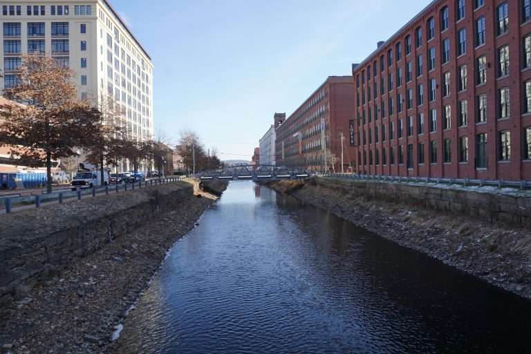 A calm canal runs between two large buildings—one white and one red brick—in Lawrence MA on a clear day, with a small bridge crossing over the water in the background. Trees and a road line the left side.