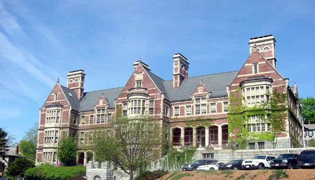 A large, historic brick building with ivy-covered walls, arched windows, and multiple chimneys, sits behind a row of trees and parked cars under a clear blue sky.