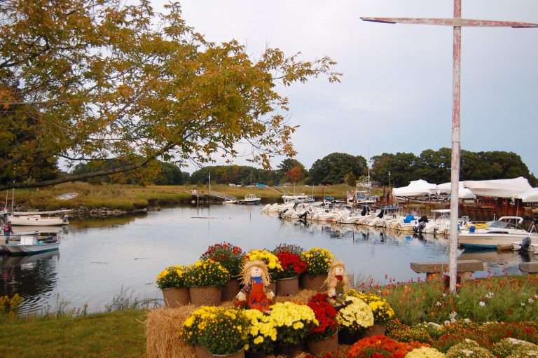 A marina with boats docked on calm water, surrounded by autumn trees. In the foreground, colorful potted flowers and two scarecrows sit on hay bales, adding a festive and rustic touch to the scene.