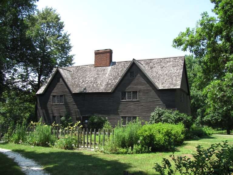 A historic dark wooden house with steep roofs, diamond-pane windows, and a red brick chimney stands surrounded by lush greenery and a simple wooden fence on a sunny day.
