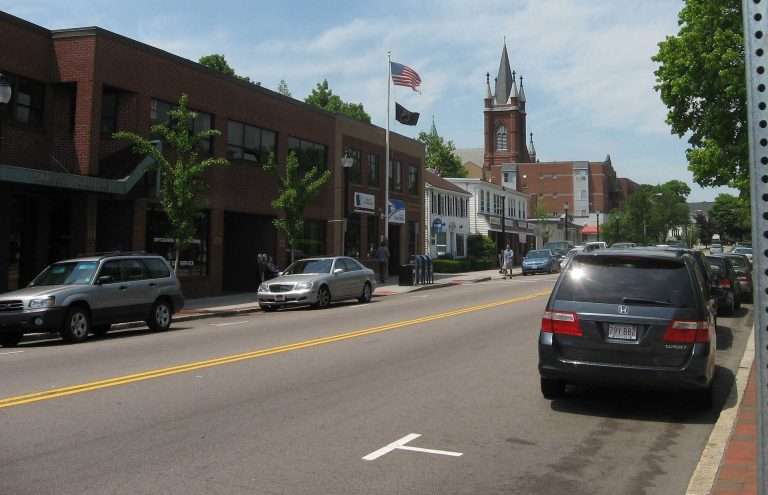 A small town street in Watertown, MA, with parked cars, brick buildings, a few trees, an American flag, and a church with a tall steeple in the background under a partly cloudy sky.