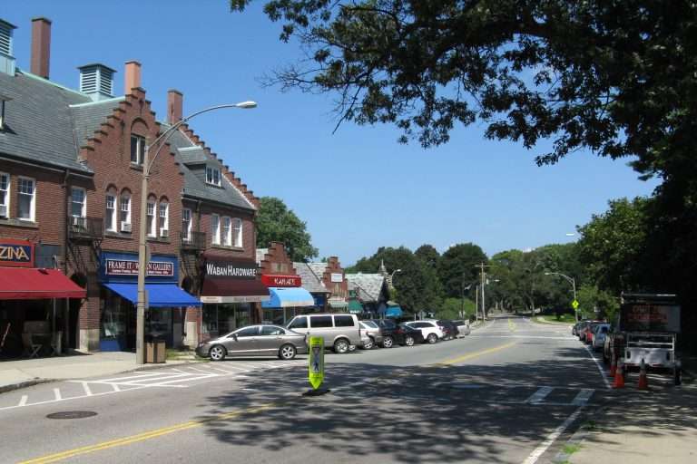 A quiet street lined with brick storefronts, awnings, and parked cars on a sunny day. Trees and a grassy park area are visible in the background under a clear blue sky.