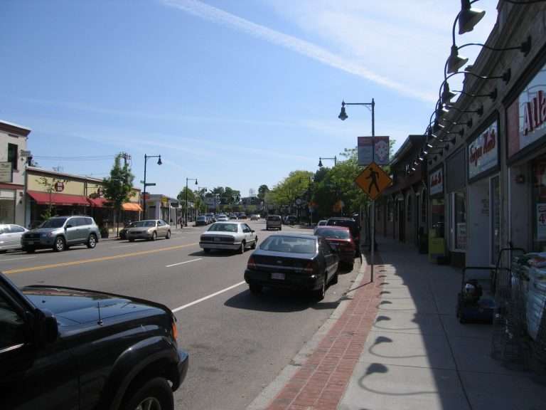A sunny day on a small town main street with parked cars, storefronts on the right, and businesses across the road. A pedestrian crossing sign is visible, along with streetlights and trees lining the street.
