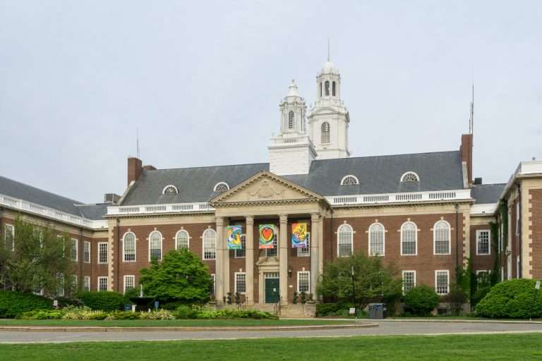 A large, historic brick building with white columns and two tall towers, featuring colorful banners above the entrance, surrounded by green trees and lawn under a cloudy sky.