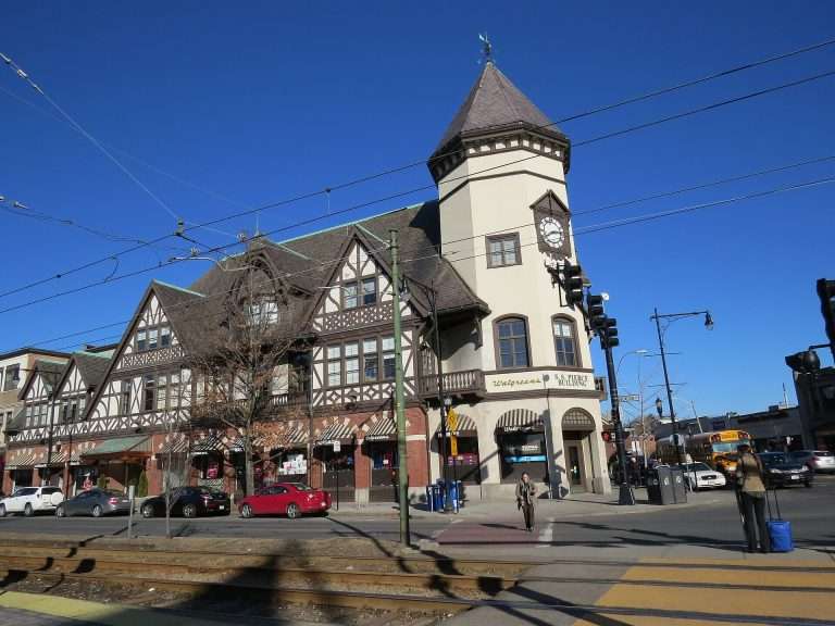 A large building with Tudor-style architecture and a clock tower stands beside a street in Brookline, MA, with cars, people, and train tracks under a clear blue sky. Shops and restaurants are located on the ground floor.