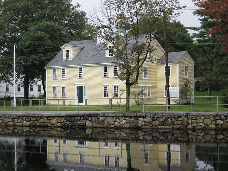 A large, yellow, colonial-style house with white trim stands behind a stone wall and calm canal in Medfield, MA, with trees and an American flag visible nearby on a cloudy day.