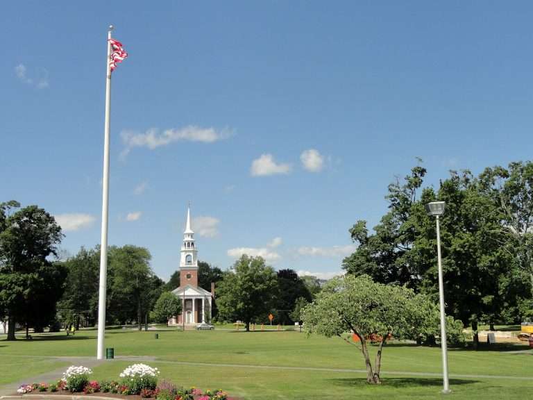 A tall flagpole with an American flag stands in a grassy Framingham, MA park with trees, flowers, and a historic white-steepled church in the background under a blue sky with scattered clouds.