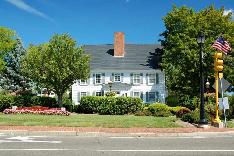 A large white colonial-style house with black shutters sits behind lush green bushes, pink flowers, and trees in Middleton MA. An American flag and streetlights are visible near a brick sidewalk under a clear blue sky.