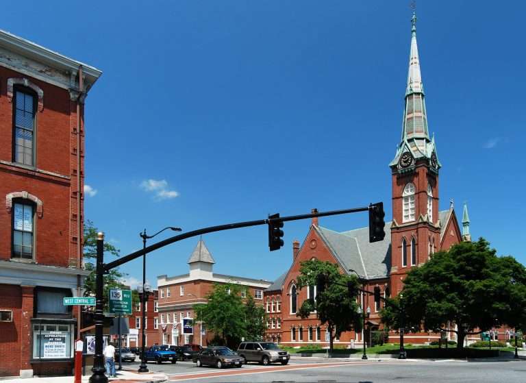 A red-brick church with a tall steeple stands on a street corner under a clear blue sky. Nearby are historic brick buildings, trees, cars, and a traffic light at the intersection of Main and Central streets.
