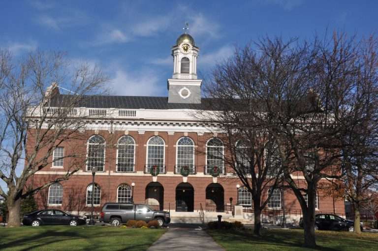 A historic red-brick building with arched windows, a white clock tower topped with a gold dome, and several cars parked in front; leafless trees frame the scene under a blue sky.