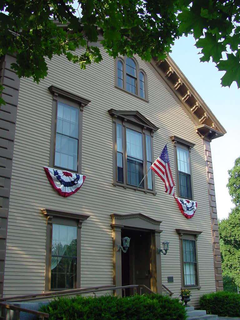A beige, two-story building decorated with American flags and red, white, and blue bunting. Green trees partially frame the top of the image, and sunlight shines on the building.