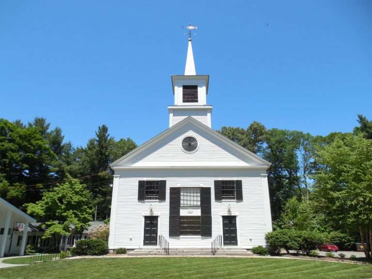 A white church with a tall steeple and black shutters stands in front of a green lawn, surrounded by trees under a clear blue sky.