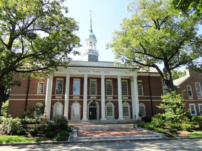 A red-brick building labeled Weston Town Hall with white columns, arched windows, and a tall cupola, surrounded by trees and greenery on a sunny day.