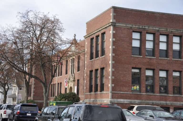 A two-story red brick school building with large windows, an arched main entrance, and an American flag stands in Wakefield, MA. Cars are parked along the street in front, and leafless trees line the sidewalk.