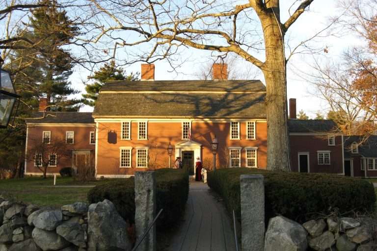 A large, historic two-story wooden house with a stone walkway, leafless tree, and surrounding shrubs, photographed in warm sunlight. A few people stand at the entrance.