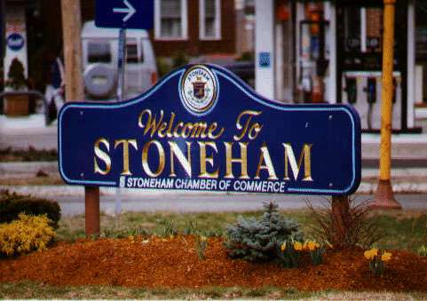 A blue sign with gold letters reads Welcome to Stoneham, MA, Stoneham Chamber of Commerce, surrounded by flowers and shrubs, with buildings and a street in the background.