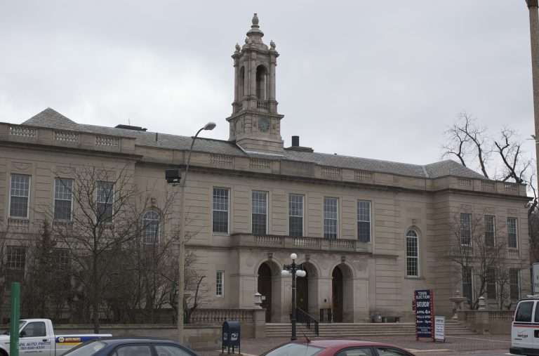 A large, historic beige stone building in Arlington, MA features tall windows, a central clock tower, and arched entrances. Seen on a cloudy day, several cars are parked in front as leafless trees line the sidewalk.