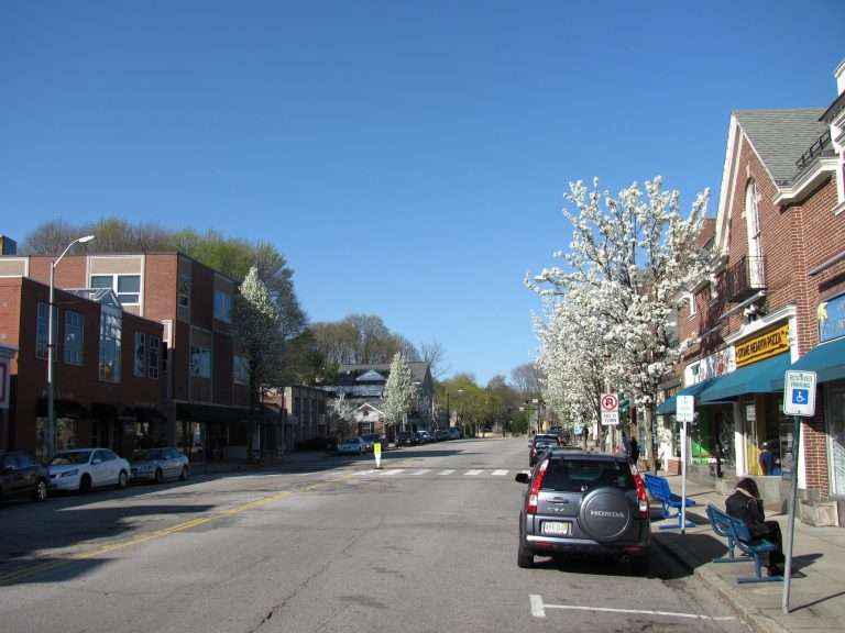 A quiet small-town street with blossoming trees, parked cars, brick and modern buildings, and a bright blue sky on a sunny day. People sit on a bench near storefronts on the right.