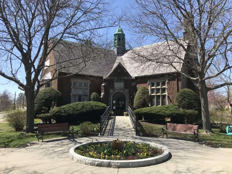 A historic brick building in Auburndale, MA, with a green cupola, large windows, and double doors, surrounded by trees, bushes, benches, and a circular flower bed in front under a clear blue sky.
