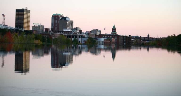 City skyline at sunset reflected in a calm body of water in Manchester, NH, with trees and various buildings, including one with a green clock tower, under a pastel sky.