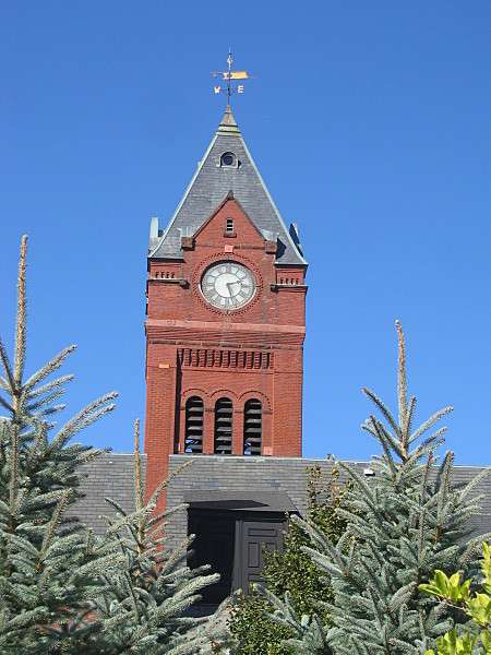 A tall red brick clock tower with a pointed gray roof rises above green trees in Winchester, MA, set against a clear blue sky.