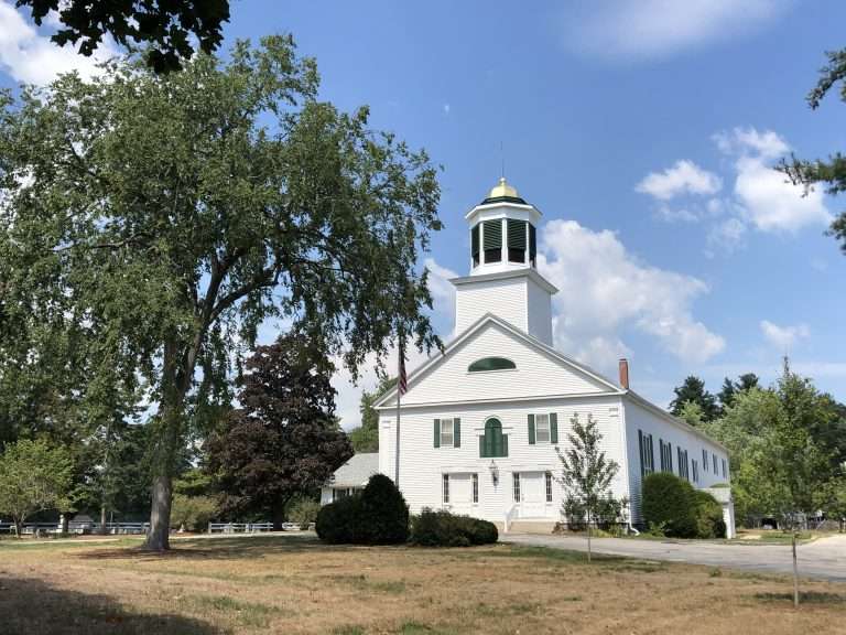 A white church with a tall steeple and green shutters stands surrounded by trees and a grassy lawn in Merrimack, NH, under a partly cloudy blue sky.