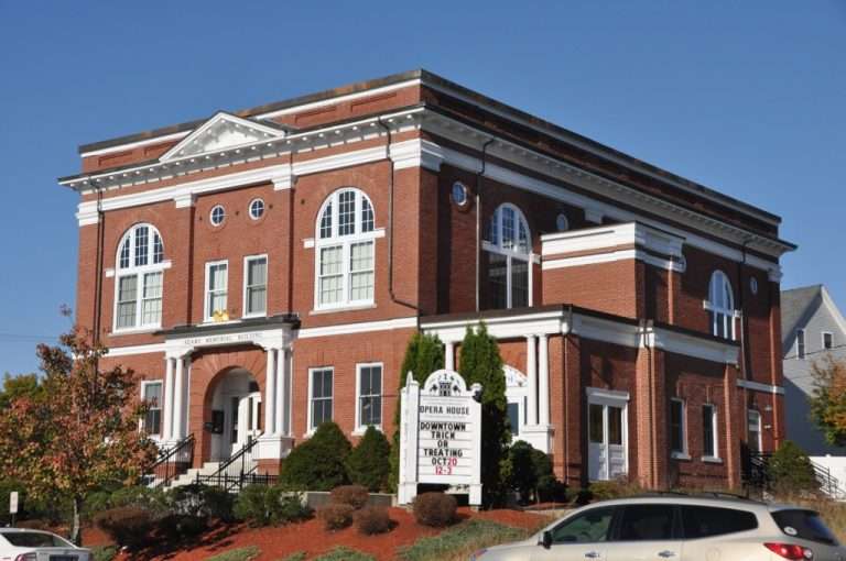 A historic red brick building with white trim and large arched windows, featuring a sign in front advertising an opera house and upcoming events, surrounded by shrubs and cars under a clear blue sky.