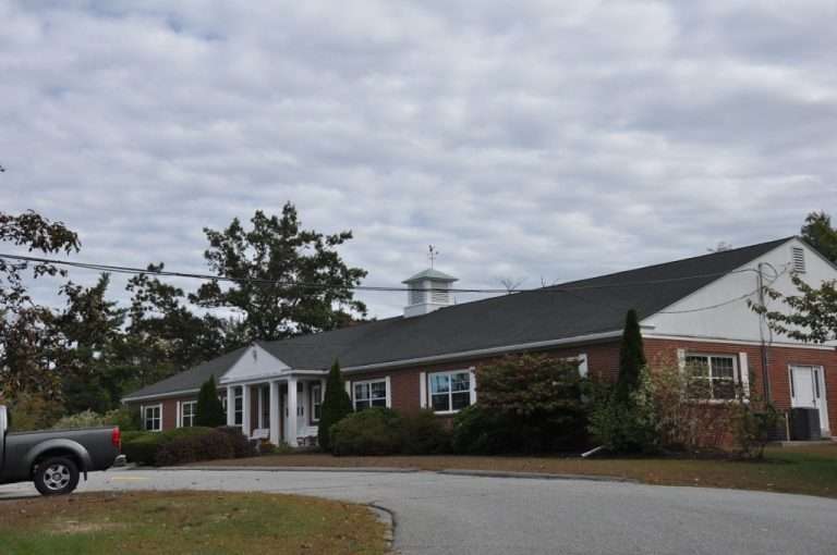A single-story red brick building with white trim and a cupola sits under a cloudy sky, surrounded by bushes and trees. A silver pickup truck is parked on a curved driveway in front of the building.