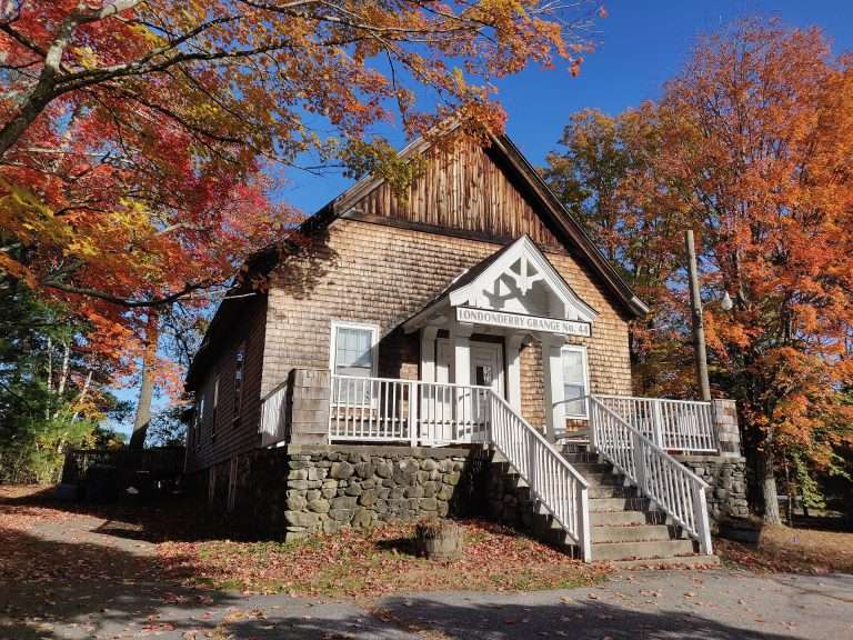 A small wooden building with a stone foundation and white-railed steps sits among colorful autumn trees in Londonderry, NH. A sign above the entrance reads Londonderry Grange No. 44 as sunlight highlights the vibrant fall foliage.