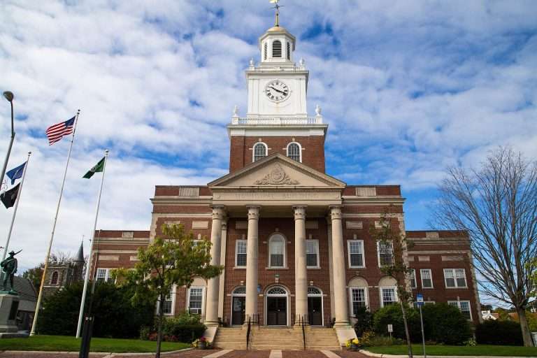 A large brick municipal building with tall columns, a clock tower, and a peaked roof stands under a partly cloudy sky. Flags are flying on poles in front, and trees line the street.