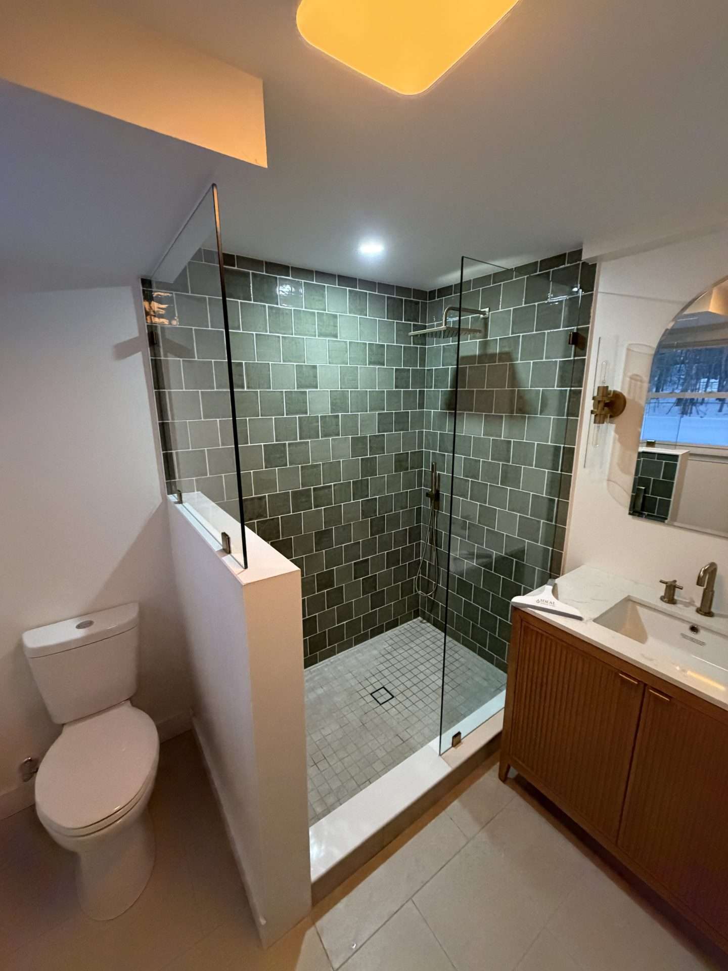 Half-wall frameless shower door with green subway tile under sloped ceiling, dark grout detail
