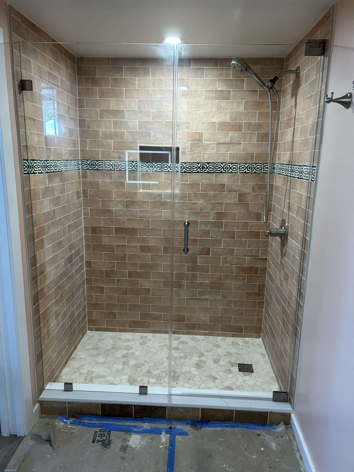 Frameless shower enclosure with warm brick tile, decorative mosaic border, natural stone floor