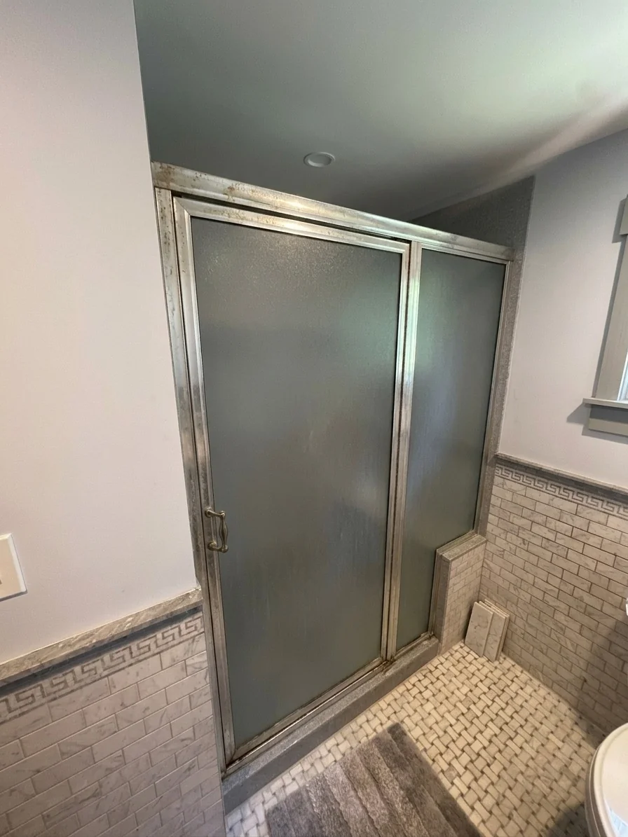 Before photo of a corroded framed sliding shower door with frosted pebbled glass panels and a deteriorated chrome header bar in a Groveland, MA bathroom with Greek key border tile